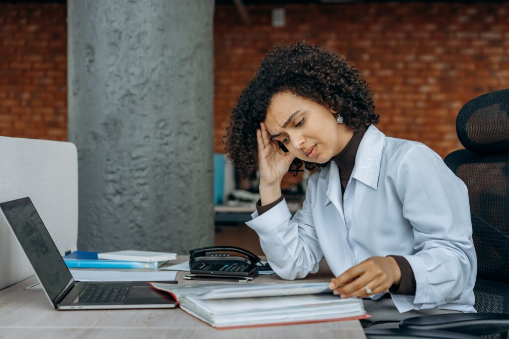 Woman at desk with computer and papers holding her head in her hand seeming tired and frustrated