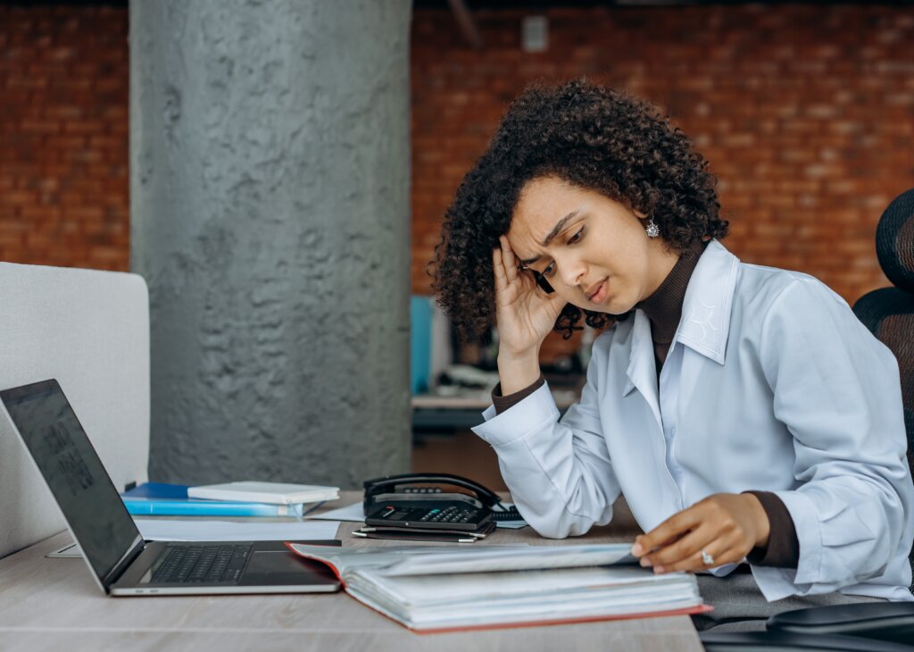 Woman at desk with computer and papers holding her head in her hand seeming tired and frustrated