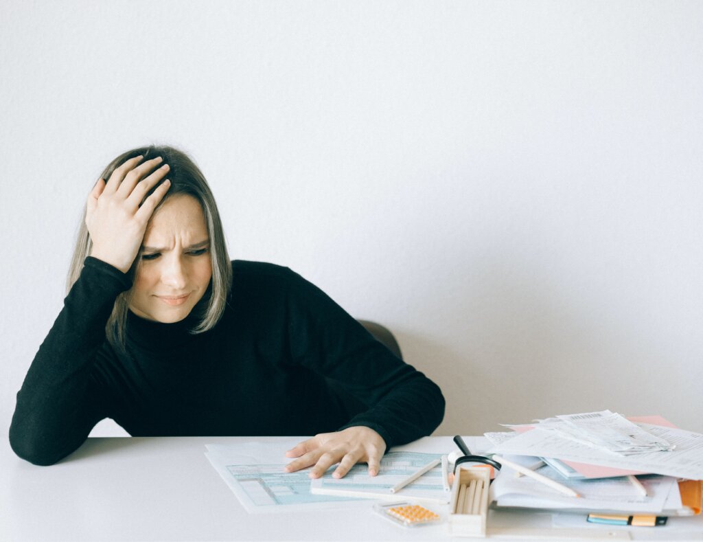 Woman in black turtleneck sitting at desk with pile of papers. She's resting her head in her hands and seems confused or frustrated