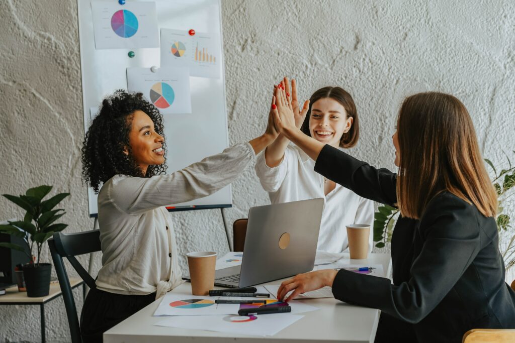Three women sitting around a laptop at a desk giving each other a high-five.