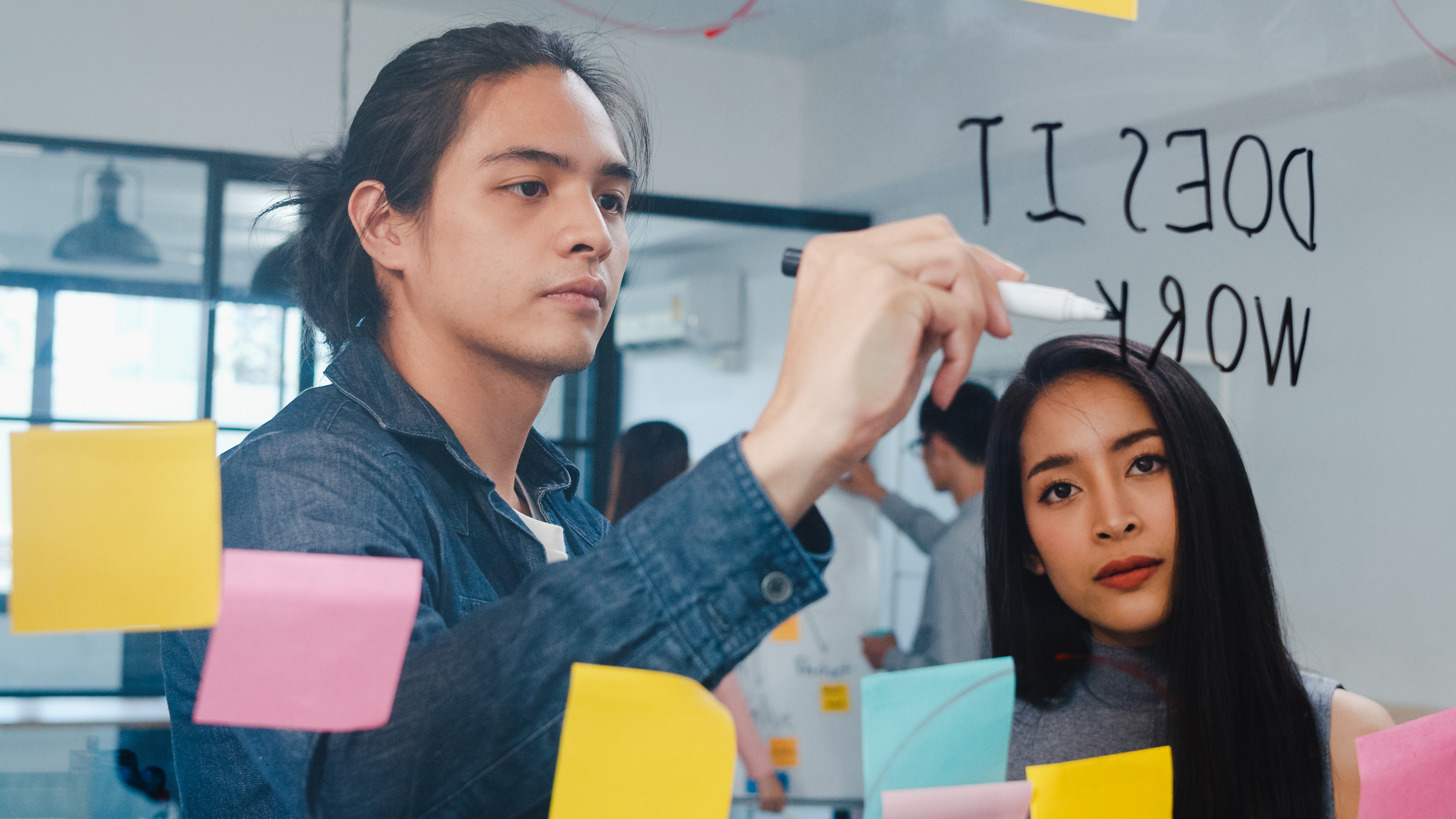 Group of young businessman and businesswoman brainstorming ideas working together sharing data and writing glass wall with sticky notes feeling happy at meeting room in small business office.