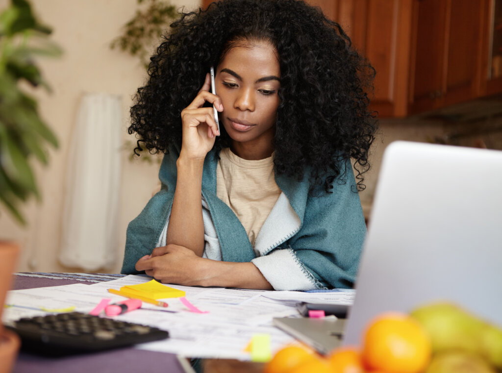Indoor shot of dark-skinned unemployed woman having phone conversation with her friend, asking her for money to pay out debts, sitting at kitchen table with laptop and documents, calculating bills