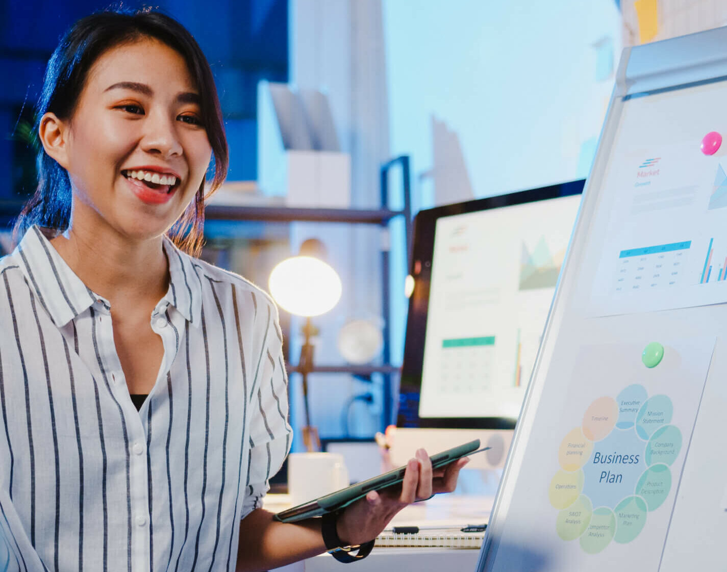 Asia businesswoman social distancing in new normal for virus prevention looking at camera presentation to colleague about plan in video call while work in office night. Lifestyle after coronavirus.