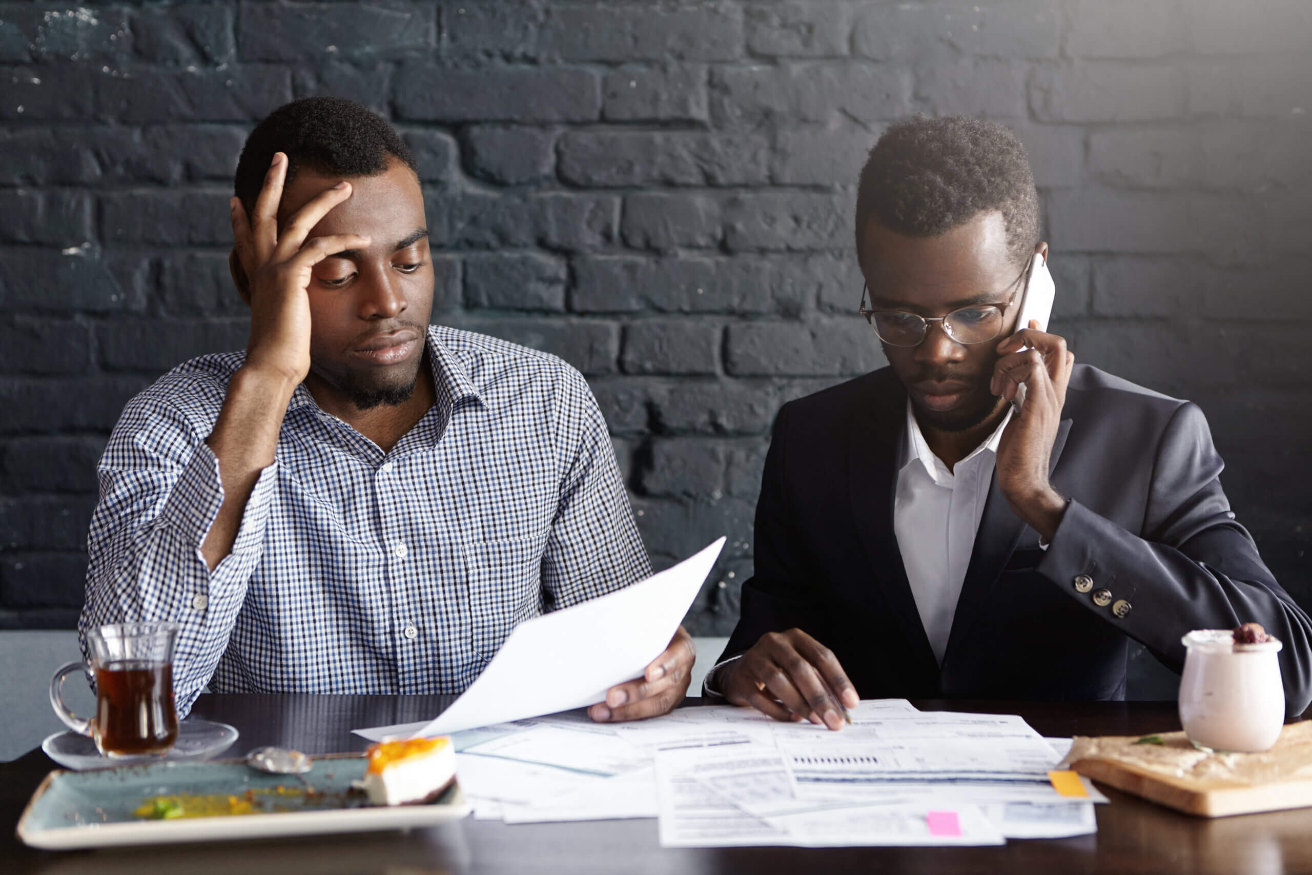 Candid shot of serious Afro-American colleagues in formal wear working together in office: man in shirt looking through papers while male wearing glasses having phone conversation, looking worried