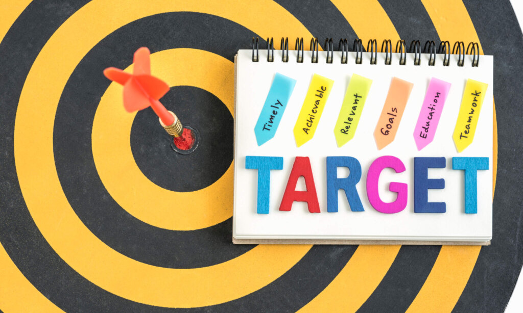 dart target in bullseye with words target on the notebook with handwriting timely achievable relevant goals education teamwork over dartboard background, Business success concept