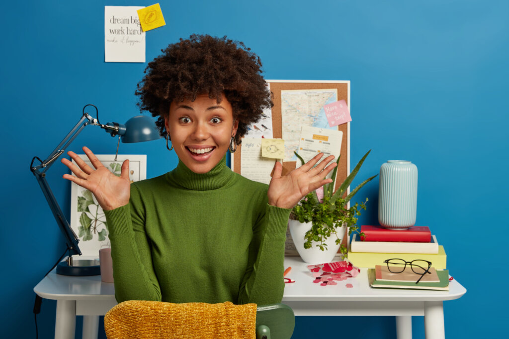 Intelligent female worker spreads palms, works in coworking space, has glad expression, dressed in green turtleneck, poses against blue background at desktop, has break after writing composition