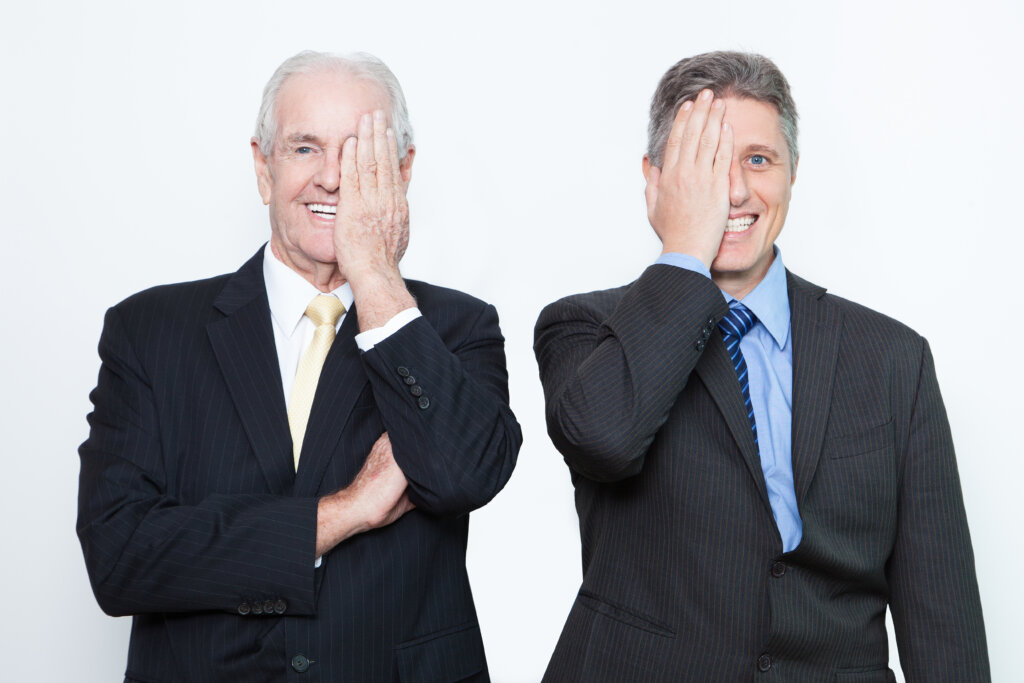 Two cheerful Caucasian businessmen examining vision and looking at camera. They closing one eye with hand. They working as team. Business concept